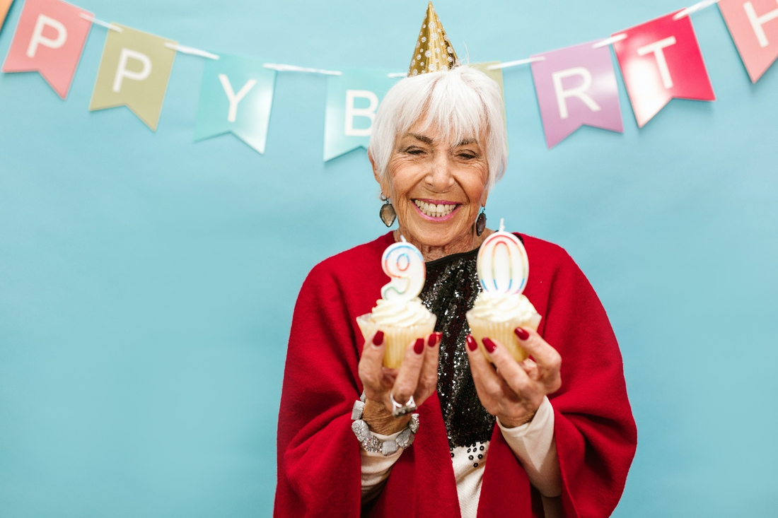 An elderly woman holding two cupcakes in front of a happy birthday banner